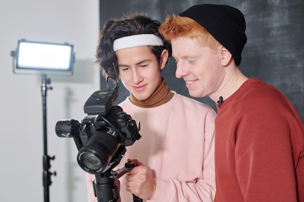 Two happy young males in casualwear watching recorded video on digital screen of camera after shooting