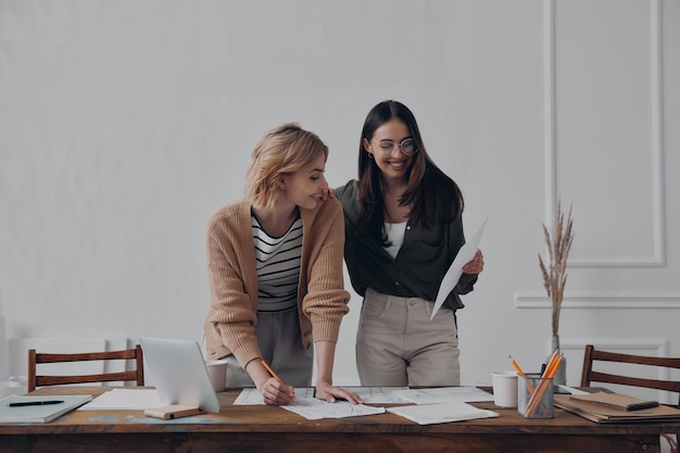 two happy young women doing paperwork while standing near the desk in office together