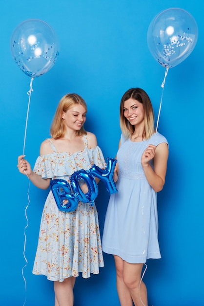 Two happy young women posing in a studio holding blue balloons and balloon with the inscription boy blue background