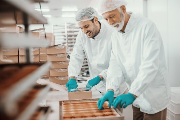 Two hardworking dedicated Caucasian employees dressed in white sterile uniforms collecting and packing cookies in boxes. Food plant interior.