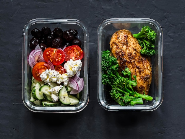 Two healthy balanced lunch boxes with greek salad baked chicken breast and broccoli on a dark