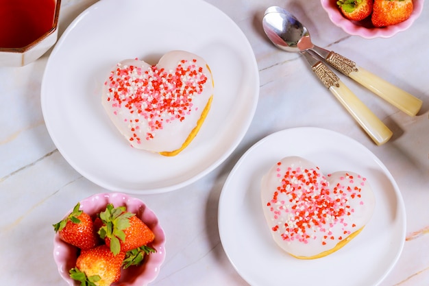 Two heart shaped donuts with white glaze and pink, red sprinkles on a platet with strawberry.