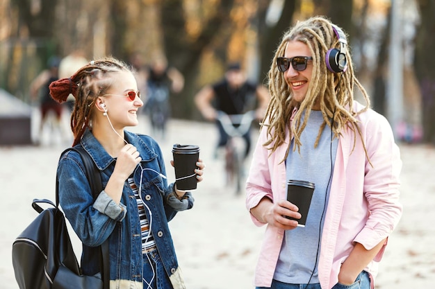 Two hipster best friends with dreads wearing headphones and glasses walking together laughing and drinking coffee in the street