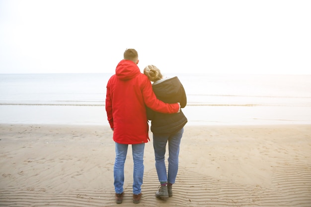 Two hipsters standing on the cold beach. Couple hugging and holding hands. Love story near the ocean. Winter season on the sea. Stylish boots on the sand. Man in red jacket.