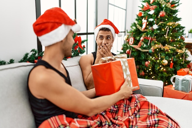 Two hispanic men couple surprise with gift sitting by christmas tree at home