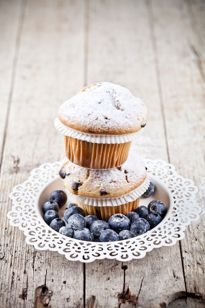 Two homemade fresh muffins with sugar powder and blueberries on white plate on rustic wooden table.