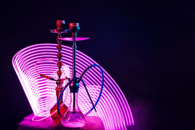 Two hookahs with pipes and bowls and a flask of water with purple neon lighting on a black background