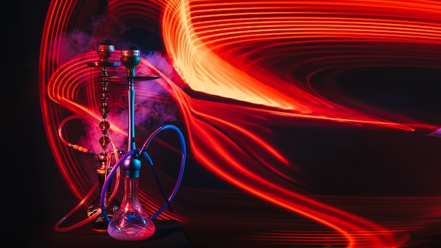 Two hookahs with shisha coals and smoke with red and blue neon lighting on the table on a dark background