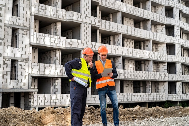 オーバーオールとヘルメットの2人の重要な労働者が、新しい建物のタブレットを見ています。建設コンセプト