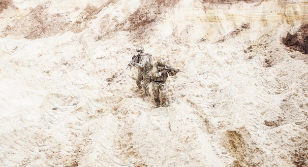 Two infantry soldiers in combat camo uniform, with tactical ammunition, carefully walking and aiming with assault rifles in unknown desert area. Military reconnaissance, scouting on enemy territory