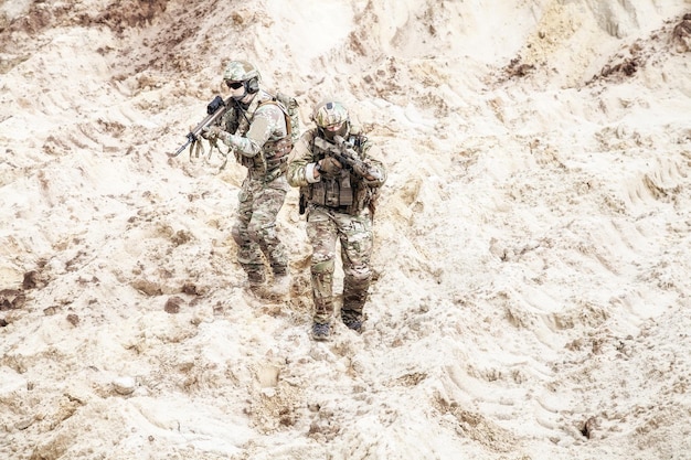 Два пехотинца в боевой камуфляжной форме с тактическими боеприпасами осторожно идут и целятся из автоматов в неизвестной пустынной местности. военная разведка, разведка территории противника