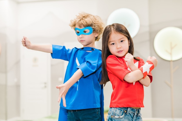 Due bambini interculturali in costumi di supereroi in piedi uno vicino all'altro davanti alla telecamera durante il gioco