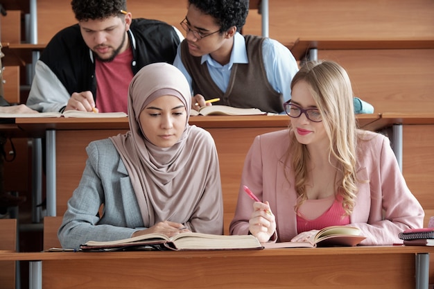 Two intercultural female students of university sitting by desk
in lecture hall and looking through passage in book during group
work at lesson