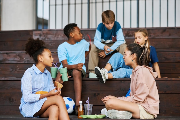 Two intercultural schoolgirls chatting while one of them having soda