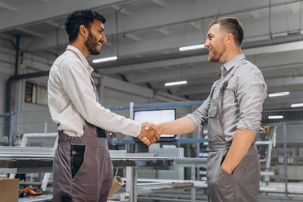 Two international male workers in overalls shake hands against the background of the production of PVC windows and doors