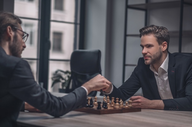 Two involved players in business suits holding hands over chessboard sitting at table in office