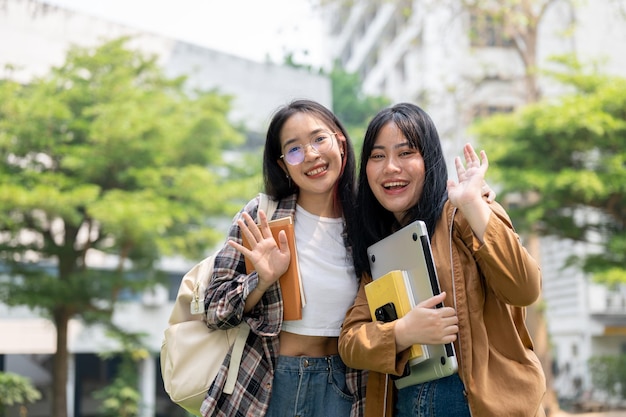 Foto due allegre studentesse universitarie asiatiche sorridono e salutano la telecamera in piedi all'aperto