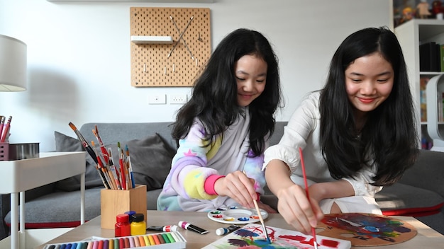 Two joyful asian painting picture together while sitting on couch at home