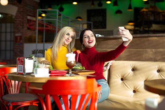 Photo two joyful cheerful girls taking a selfie while sitting together at cafe