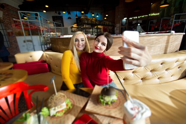 Two joyful cheerful girls taking a selfie while sitting together at cafe