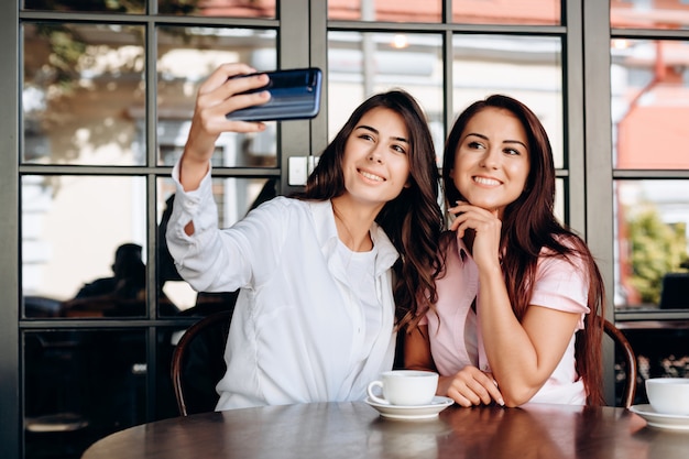 Two joyful cheerful girls taking a selfie while sitting together at cafe