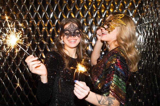 Photo two joyful girls in venetian masks holding sparkling bengal lights while having fun at party in the night club