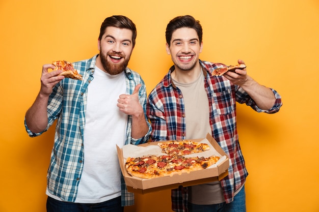 Two joyful men holding pizza  while bearded man showing thumb up over yellow wall