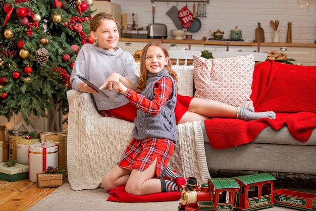 즐거운 두 자매가 집에 크리스마스 트리 근처에 앉아 태블릿 모니터를 보고 있습니다