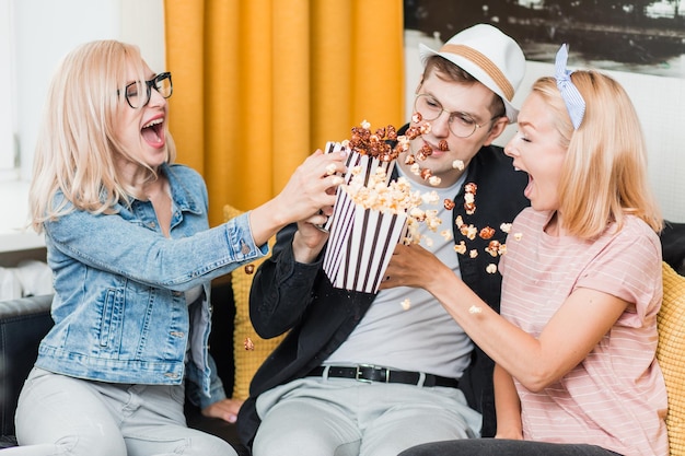 Two joyful young caucasian blonde women and man having fun, eating popcorn and enjoying together. Cheerful group of friends sitting on couch watching TV show and celebrating at home.