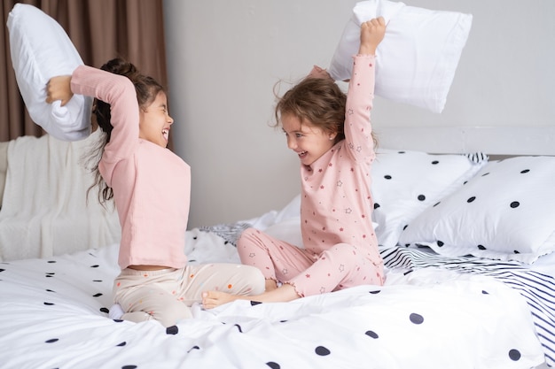 Two kids girls in pajamas having pillow fight on bed in modern bright apartment