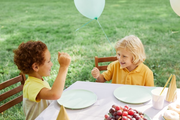 Two kids at picnic table outdoors decorated with balloons for birthday party in summer copy space