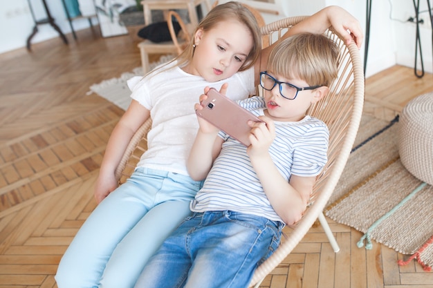 Two kids playing smartphone. Boy and girl at home watching at the cell phone. Digital children indoor.