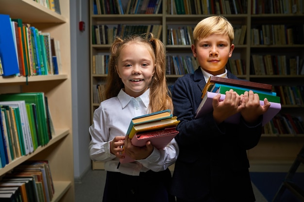 two kids prepared for school, they stand holding stack of books in hands, looking at camera, wearing schools outfit
