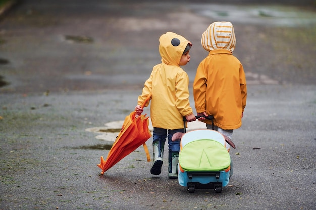 Two kids with umbrella suitcase and yellow waterproof cloaks and boots walking outdoors after the rain together