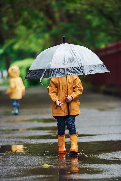 Two kids with umbrella in yellow waterproof cloaks and boots playing outdoors after the rain together