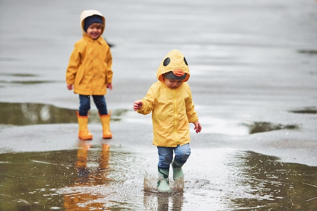 一緒に雨の後に屋外で遊ぶ黄色の防水マントとブーツの2人の子供