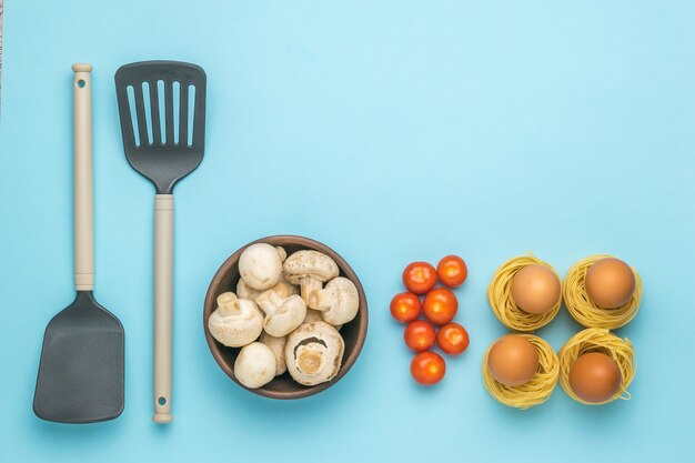 Due spatole da cucina, pasta, uova, funghi e pomodori su sfondo blu. ingredienti per fare la pasta. Foto Premium
