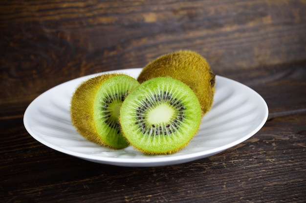 Two kiwi fruits on white plate, one kiwi cut in half, still life on brown wooden plank background.