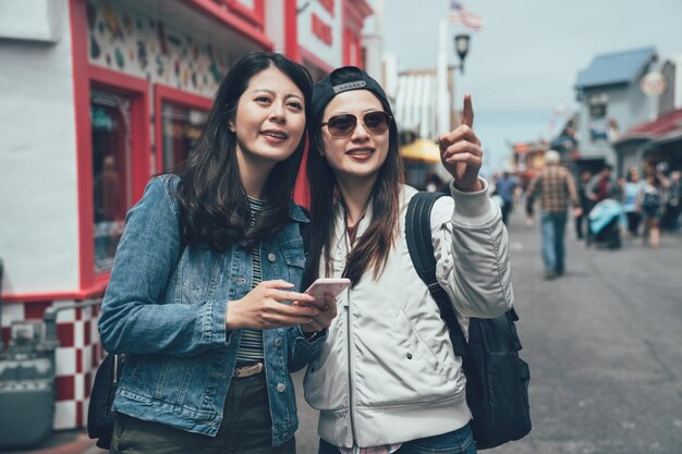 Two korean young girl travelers holding mobile phone online map\
consider right way next destination standing outdoor weekend\
farmers market in old fisherman\'s wharf monterey on holiday.\
teeming place