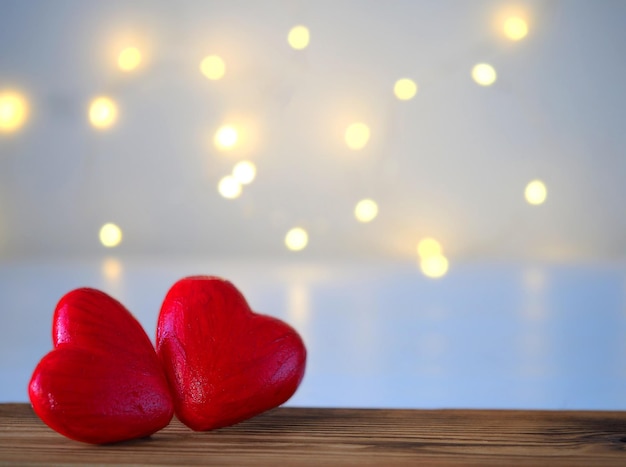 Two large red hearts lie on a wooden table on a blur compared background with a side from the garland, a horizontal image with a soft focus and a place for text