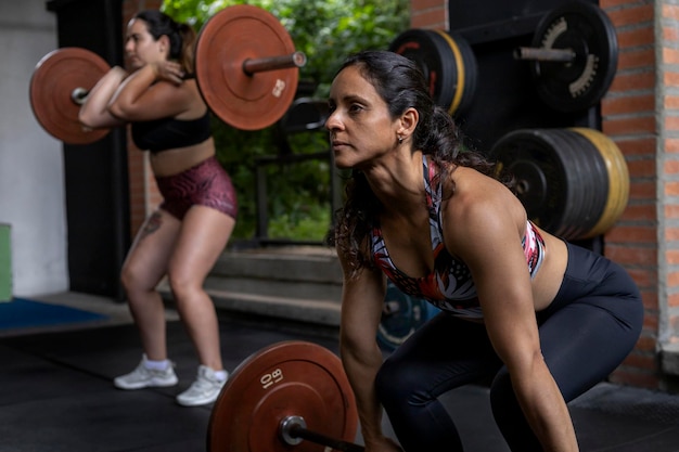 두 명의 라틴 아메리카 여성이 웨이트가 있는 바벨을 사용하여 체육관에서 고강도로 실행되는 기능적 운동을 수행합니다.