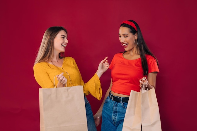 Two laughing dancing young sisters girls in bright clothes are\
holding a package with purchases isolated on a red background
