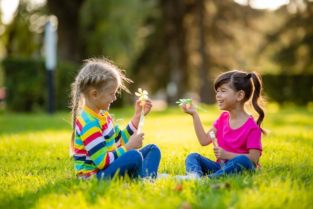 2人の小さなブロンドの女の子とブルネットの夏は、シャボン玉を吹く芝生の上に座っていますヨーロッパとインドの民族の子供たち