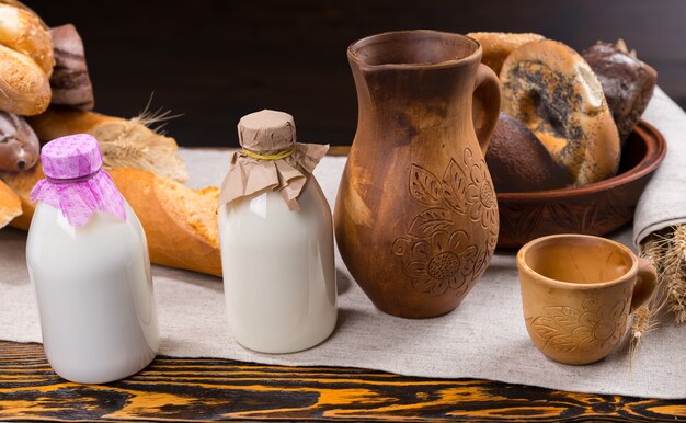 Two little bottles of milk with paper covers next to wooden pitcher, cup and various loaves and rolls of bread on table