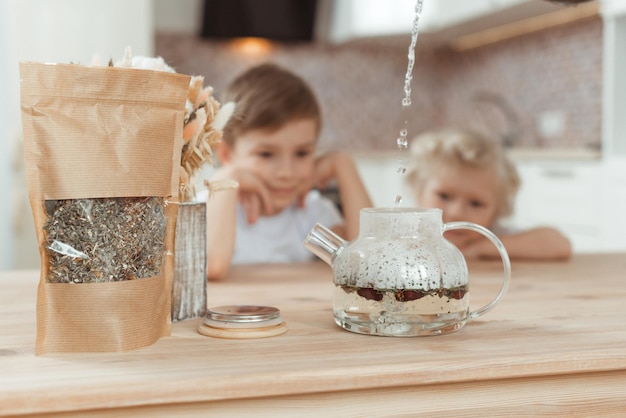 Two little boys are sitting at the kitchen table watching water pour into a teapot