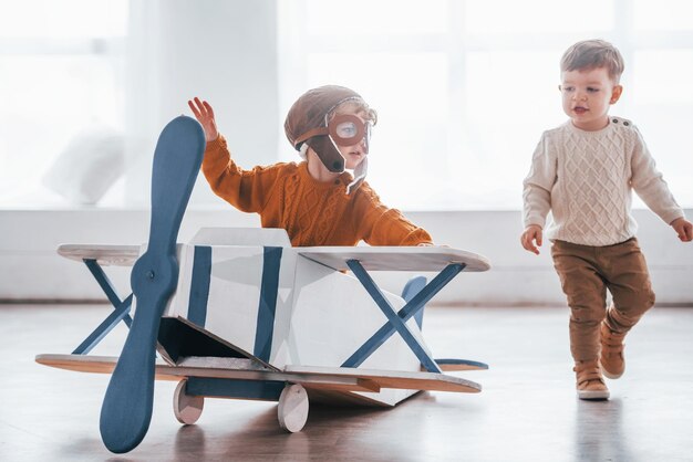 Two little boys in retro pilot uniform having fun with toy plane indoors