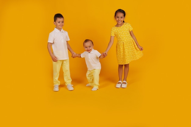 Two little boys in yellow trousers and a white tshirt hold the hand of a sister in a yellow dress