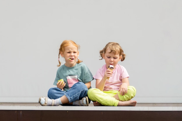 2人の小さな白人のかわいい女の子が足を組んで座ってアイスクリームを食べています。