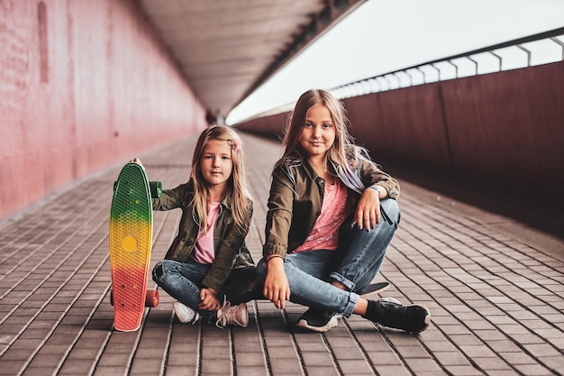 Two little cheerful sisters are sitting on the skateboard in the tunnel.