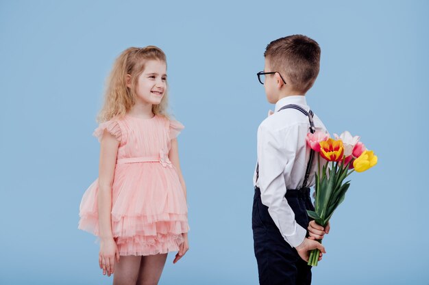 Two little children boy with flowers and girl in pink dress, isolated on blue wall